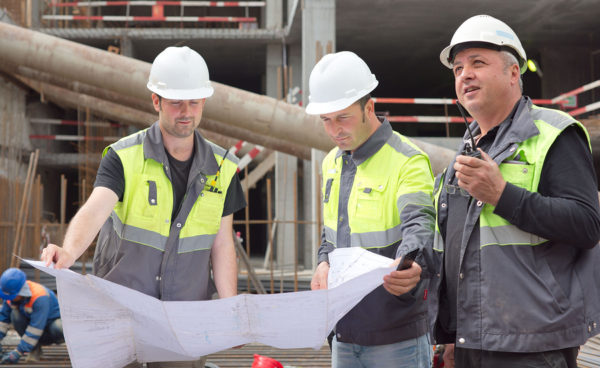 Two civil engineers and a senior foreman at construction site are inspecting ongoing construction works according to design drawings.